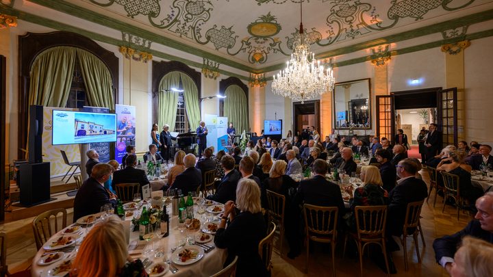 During the charity gala in Kittsee, a large group of people in formal attire sits at round tables in a lavishly decorated hall, listening to the speakers on stage. A magnificent chandelier hangs from an elaborately decorated ceiling. 