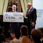 At the charity gala in Kittsee, two men in suits are presenting a large check for 10,000 euros during an event in the hall, while a woman stands next to them holding a microphone. A banner and a piano can be seen in the background. 