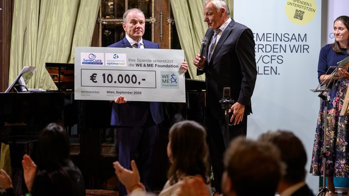 At the charity gala in Kittsee, two men in suits are presenting a large check for 10,000 euros during an event in the hall, while a woman stands next to them holding a microphone. A banner and a piano can be seen in the background. 