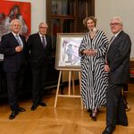 During the charity gala in Kittsee, four people dressed in evening attire are standing in a gallery around a framed piece of art; abstract paintings hang on the walls, and bottles of wine sit on a side table. The setting is elegant, with wooden floors and classic furnishings. 