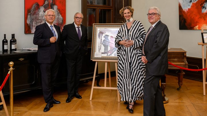 During the charity gala in Kittsee, four people dressed in evening attire are standing in a gallery around a framed piece of art; abstract paintings hang on the walls, and bottles of wine sit on a side table. The setting is elegant, with wooden floors and classic furnishings. 