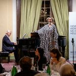 A woman in a black-and-white striped dress is singing next to a pianist at a festive event in a hall, presumably as part of the Kittsee Charity Gala, while the audience sits at tables and two large banners are visible behind the performers.