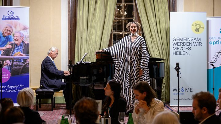 A woman in a black-and-white striped dress is singing next to a pianist at a festive event in a hall, presumably as part of the Kittsee Charity Gala, while the audience sits at tables and two large banners are visible behind the performers.