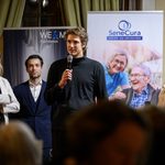 A man is speaking into a microphone while three women and a man stand next to him at the charity gala in Kittsee; in the background, a SeneCura banner featuring smiling seniors can be seen.