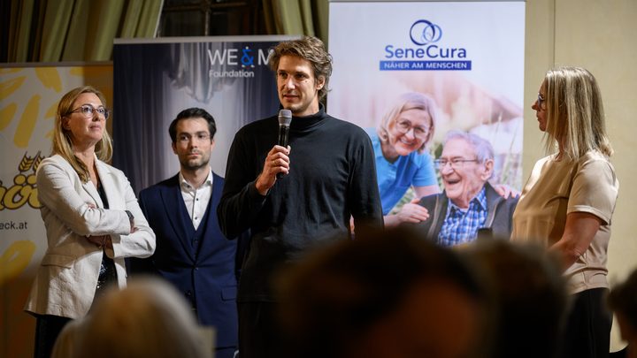 A man is speaking into a microphone while three women and a man stand next to him at the charity gala in Kittsee; in the background, a SeneCura banner featuring smiling seniors can be seen.