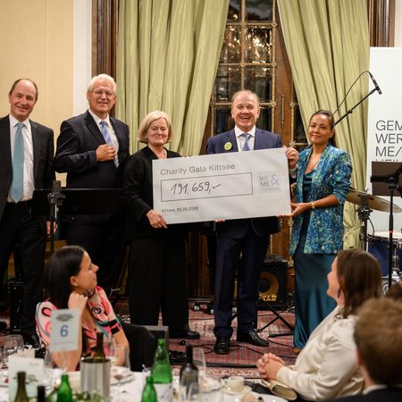 Six people are standing at the charity gala in Kittsee, holding a large check. The hall is elegantly decorated; in the background, guests are seated, and musical instruments and banners for SeneCura and ME/CFS are visible. The atmosphere is festive.  