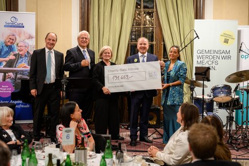 Image of Six people are standing at the charity gala in Kittsee, holding a large check. The hall is elegantly decorated; in the background, guests are seated, and musical instruments and banners for SeneCura and ME/CFS are visible. The atmosphere is festive.  