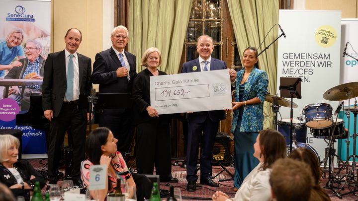 Six people are standing at the charity gala in Kittsee, holding a large check. The hall is elegantly decorated; in the background, guests are seated, and musical instruments and banners for SeneCura and ME/CFS are visible. The atmosphere is festive.  